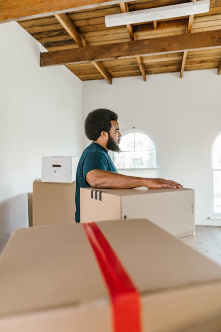 A man with dark curly hair and a beard, wearing a teal shirt, is seen in an indoor space with a high wooden ceiling and white walls, preparing for a home relocation. He is standing behind a stack of l